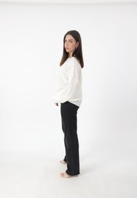 Witte kabelgebreide trui met een ontspannen pasvorm, gecombineerd met zwarte flared jeans en lichtgekleurde haksandalen. Model staat voor een witte achtergrond.