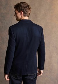 Man med brunt hår som bär en mörkblå kavaj och mörka jeans, stående vänd åt sidan mot en enkel texturerad bakgrund.