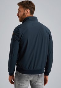 Marineblaues Bomberjacke mit hohem Kragen, weichem Stoff, gerippten Manschetten und einem strukturierten Akzent auf der Schulter. Graue Jeans sind darunter sichtbar.