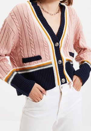 Vrouw draagt een roze en marineblauwe kabelgebreide cardigan met gestreepte rand, zilveren knopen en witte broek, handen in de zakken.