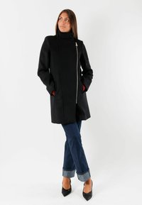 Cappotto in misto lana nero con colletto alto, chiusura con zip argentata, tasche laterali con foderatura rossa e taglio dritto, abbinato a jeans blu svasati.