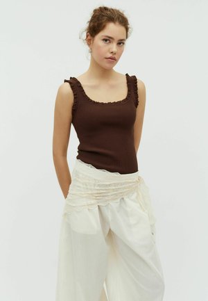 Ung kvinde med krøllet hår iført en brun, ærmeløs ribstrikket top med flæsekant og løse hvide bukser med bælte i blonder.