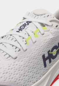 Λευκό αθλητικό παπούτσι από δίχτυ με λευκά κορδόνια, κίτρινα στοιχεία, μαύρα λογότυπα HOKA και παχύ πάτο με κόκκινες λεπτομέρειες.