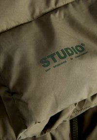 Detailní záběr na olivově zelenou polstrovanou tkaninu s tmavozeleným textem "STUDIO" a menším potiskem "ZALOŽENO V DVACETI DVACETI".