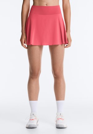 Frau trägt einen korallenrosa sportlichen Skort mit weißen Knöchelsocken und weißen Sneakers, gezeigt von der Taille bis zu den Füßen vor weißem Hintergrund.