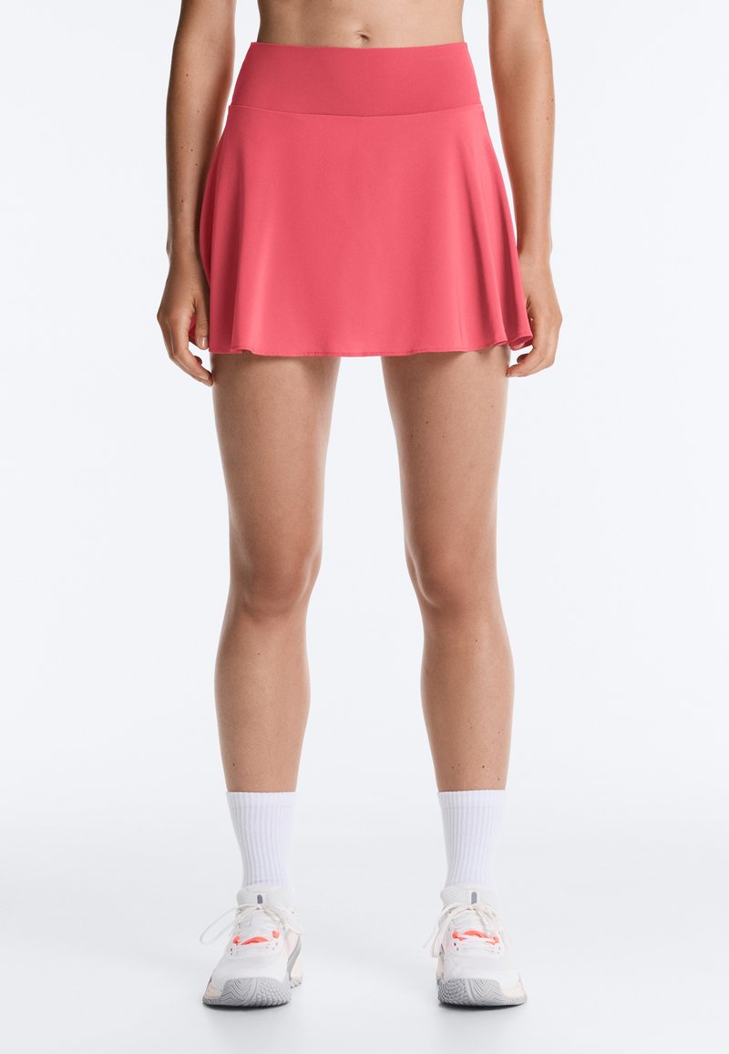 Femme portant un skort de sport rose corail avec des chaussettes blanches aux chevilles et des baskets blanches, visible de la taille aux pieds sur un fond blanc.