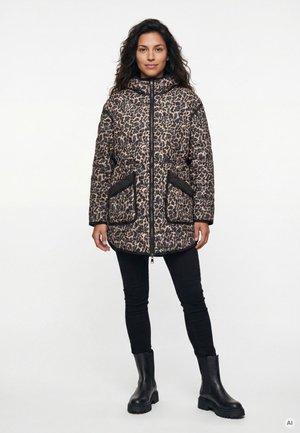 Frau trägt eine gesteppte Jacke mit Leopardenmuster, großen schwarzen Taschen, schwarze Hose und schwarze Stiefeletten und steht vor einem einfarbigen Hintergrund.