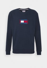 Mörkblå sweatshirt med V-hals, prydd med en röd och vit broderad logotyp på bröstet samt ribbade muddar och fåll. Mjuk, slät textur.