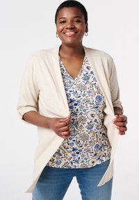 Cardigan châle crème sur un haut à motifs floraux bleus, avec un décolleté en V et un jean bleu. Tissu léger, manches trois-quarts.