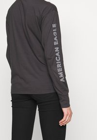 Dlouhý rukáv, tmavě šedé tričko z bavlny. Na levém rukávu vertikálně bílé písmo "AMERICAN EAGLE". Měkká textura, volný střih.
