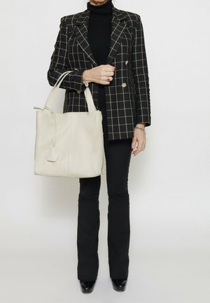 Borsa tote in pelle beige con forma strutturata, dotata di due manici e un accento esagonale sul lato. Indossata con un blazer a scacchi nero.