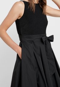 Czarna sukienka z dopasowanym, błyszczącym topem i rozkloszowaną, teksturowaną spódnicą. Zawiera akcent w formie kokardy w talii oraz kieszenie po obu stronach.