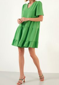 Femme portant une robe verte ample à volants avec des manches courtes bouffantes et des sandales à talons orange avec des lanières décoratives.