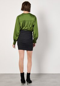 Grüne Satinbluse mit gerafftem Rücken, kombiniert mit einem schwarzen gerafften Minikleid. Das Model trägt schwarze Knöchelstiefel und posiert von hinten.