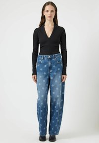 Černý top s dlouhým rukávem a výstřihem do V v kombinaci s modrými džínami s velkým puntíkovaným vzorem a volným střihem. Černé kotníkové boty.