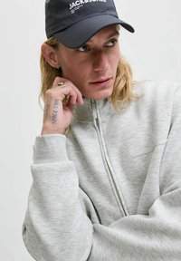 Ung person med langt blondt hår iført en sort Jack & Jones kasket og en lys grå zip-up sweatshirt, hviler hagen på en tatoveret hånd.