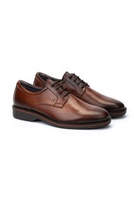 Chaussures habillées en cuir marron avec une texture lisse, bout rond, design à lacets et une marque discrète. Elles possèdent une fine semelle noire avec des accents rouges.