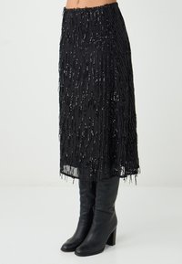 Gonna nera con frange caratterizzata da una finitura testurizzata e paillettes sparse. Abbinata a stivali neri alti fino al ginocchio e con orlo dritto.