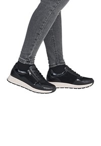 Schwarze Sportschuhe mit glatter Textur, ausgestattet mit einem glänzenden Akzent, Schnürung auf der Vorderseite und einer kontrastierenden weißen Sohle. Getragen mit grauen Jeans.