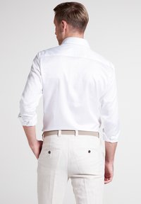 Camicia bianca con maniche arrotolate, vista da dietro. Abbinata a pantaloni beige chiaro e una cintura intrecciata. Texture liscia, design minimalista.