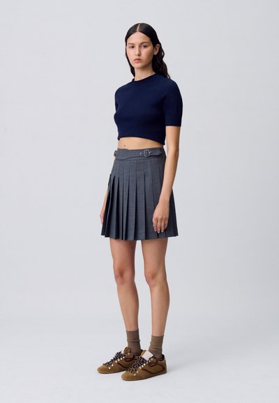 Suéter recortado azul marino, falda plisada gris con cinturón y zapatillas marrones con cordones oscuros. La modelo se encuentra en un fondo neutro.
