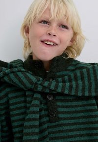 Sudadera de lana a rayas verdes y verde oscuro con cuello alto y botones negros, con un detalle de bufanda cruzada en el escote.