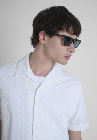 Camicia bianca a maniche corte con pattern a diamante testurizzato, colletto classico e bottoni. Gli occhiali da sole neri di forma rettangolare completano l'aspetto.