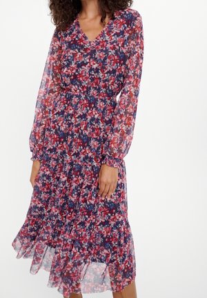 Donna che indossa un vestito floreale al ginocchio con maniche lunghe trasparenti e vita stretta in tonalità di rosso, rosa e blu su sfondo bianco.