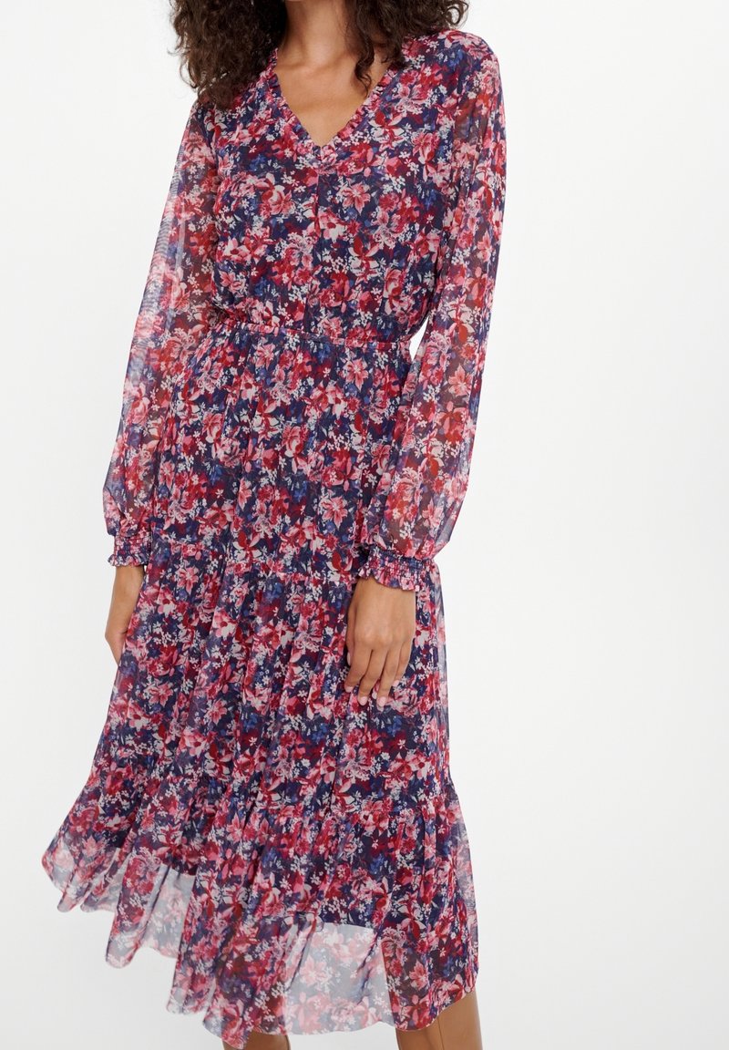 Femme portant une robe fleurie longueur genou avec manches longues transparentes et taille cintrée dans des tons de rouge, rose et bleu sur fond blanc.