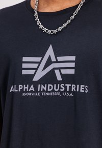 Mørkeblå bomulds T-shirt med et stort grått grafisk logo med stiliserede vinger og teksten: "ALPHA INDUSTRIES, KNOXVILLE, TENNESSEE, U.S.A."