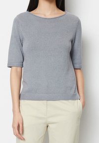 Kobieta w jasnoszarej, krótkiej bluzce dzianinowej i beżowych spodniach, z jedną ręką w kieszeni, na plain białym tle.