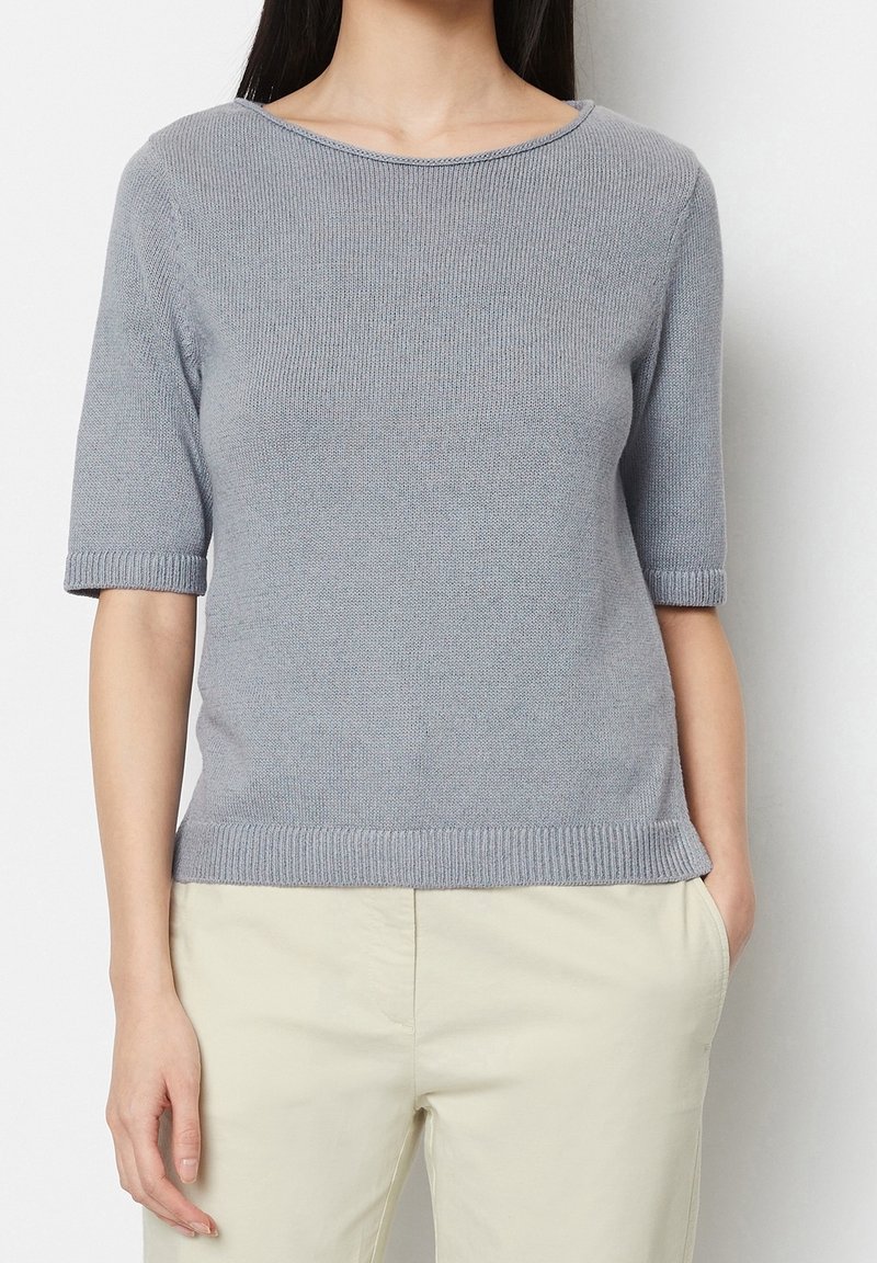 Kobieta w jasnoszarej, krótkiej bluzce dzianinowej i beżowych spodniach, z jedną ręką w kieszeni, na plain białym tle.