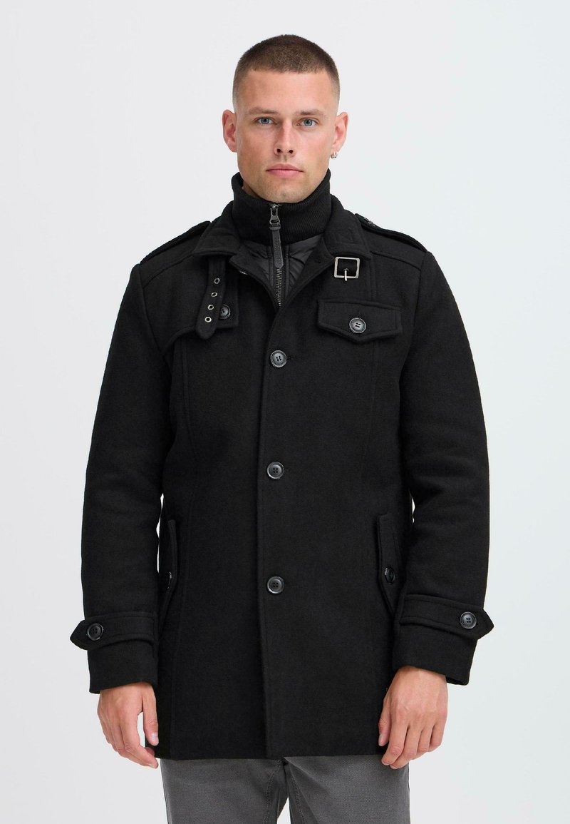 Schwarzer Wollmantel mit hohem Kragen, geknöpftem Vorderteil und mehreren Taschen. Verfügt über eine zusätzliche gefütterte Innenlage und dekorative Beschläge.