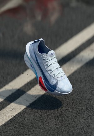 White and blue running shoe with red sole detail floating above asphalt road with white line markings.