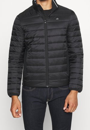 Mężczyzna ubrany w czarną pikowaną kurtkę zapinaną na zamek marki Calvin Klein z elastycznymi mankietami oraz ciemne dżinsy, widziany od szyi do bioder.