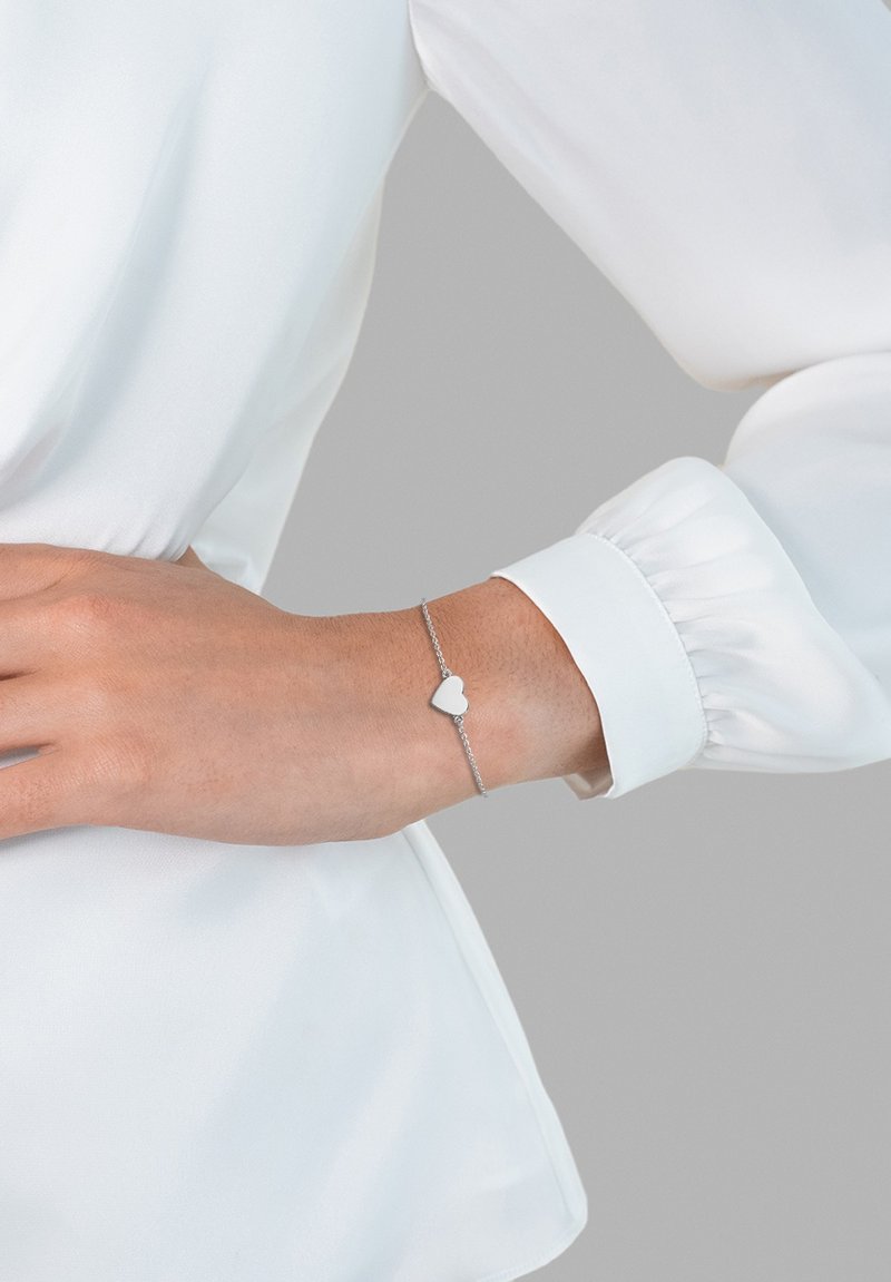 Bracelet en argent avec un pendentif en forme de cœur, affiché sur un poignet, sur fond gris clair, porté avec un blouse blanche.