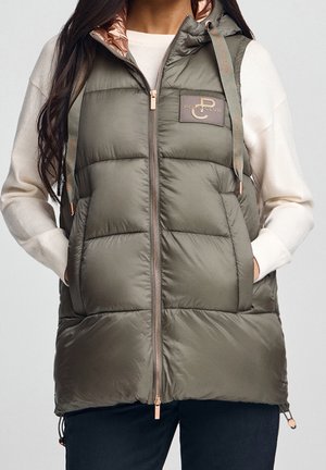 Donna che indossa un gilet imbottito verde oliva con cappuccio, cerniera color oro rosa e logo del polo club, sopra un maglione bianco a maniche lunghe con le mani nelle tasche.