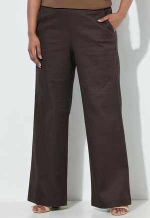 Person trägt weit geschnittene dunkelbraune Hose, beigefarbenes Oberteil und offene Schuhe, steht mit einer Hand in der Tasche.