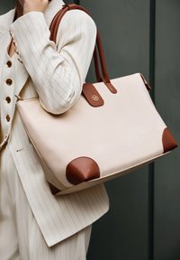 Beige læder tote taske med brune læderdetaljer ved hjørnerne, lynlås lukning og mærke logo detaljer på fronten. Stribet baggrund.