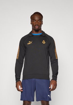 Sweatshirt noir à manches longues avec col, présentant des logos dorés et des accents à motifs sur les manches, associé à un short bleu marine.