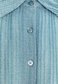 Jasnoniebieska, teksturowana tkanina o błyszczącym wykończeniu. Posiada zapięcia na guziki i subtelne fałdy w okolicy kołnierza.