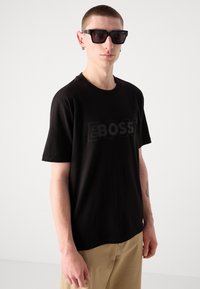 Juodas trumpas rankovės marškinėliai su reljefiškai iškilusiu „BOSS“ logotipu. Modelis dėvi masyvias juodas saulės akinius. Šviesiai rudos kelnės užbaigia aprangą.
