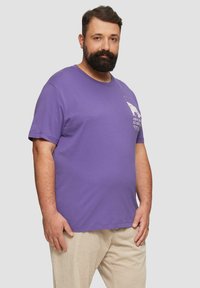Purple cotton t-shirt with short sleeves, featuring a graphic print on the chest. Smooth texture and relaxed fit. Paired with beige corduroy pants.