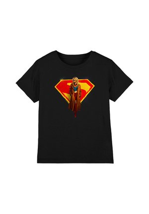 Schwarzes T-Shirt mit kurzen Ärmeln, das eine Frau in einem braunen Mantel und roten Stiefeln zeigt, die vor einem großen roten und gelben Superman-Emblem steht.