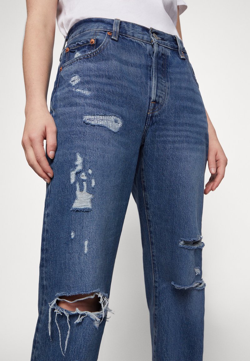 Jeans de mezclilla azul claro con cintura alta, que presentan detalles desgastados y dobladillos deshilachados. Botones de cobre acentúan la cintura y los bolsillos.