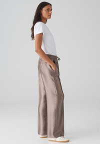 Lichte witte t-shirt en losvallende, glanzende taupe broek met elastische taille en zijzakken, gedragen met witte sneakers.
