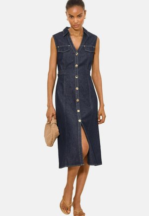 Žena nosi tmavé, bezrukávové midi džínové šaty s prednými gombíkmi, náprsnými vreckami a predným rozparkom, drží pletenú hnedú kabelku.