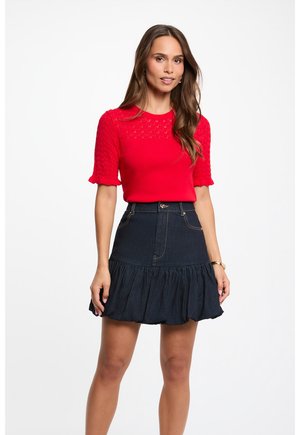 Kvinde iført en rød kortærmet strikket top med blonde detaljer og et højtaljet mørkeblåt voluminøst denimskørt.