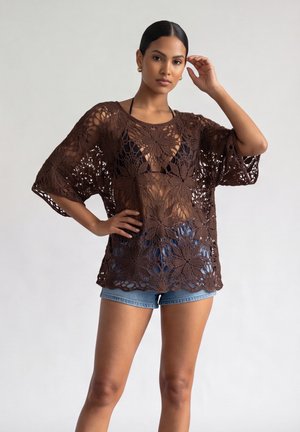 Donna che indossa un top marrone all'uncinetto sopra un bikini nero e shorts di jeans blu, in piedi con una mano sul fianco e l'altra vicino alla testa.