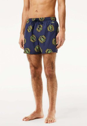 Bañadores de natación en color navy con cintura elástica y cordón, que presentan un logo de cocodrilo verde repetido y acentos de texto en forma circular. Textura suave.